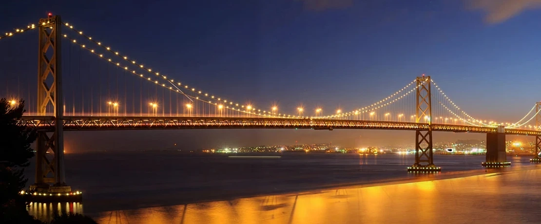 The Oakland Bay Bridge at night, beautifully lit with golden lights reflecting on the water.