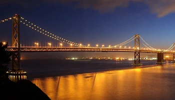 The Oakland Bay Bridge at night, beautifully lit with golden lights reflecting on the water.