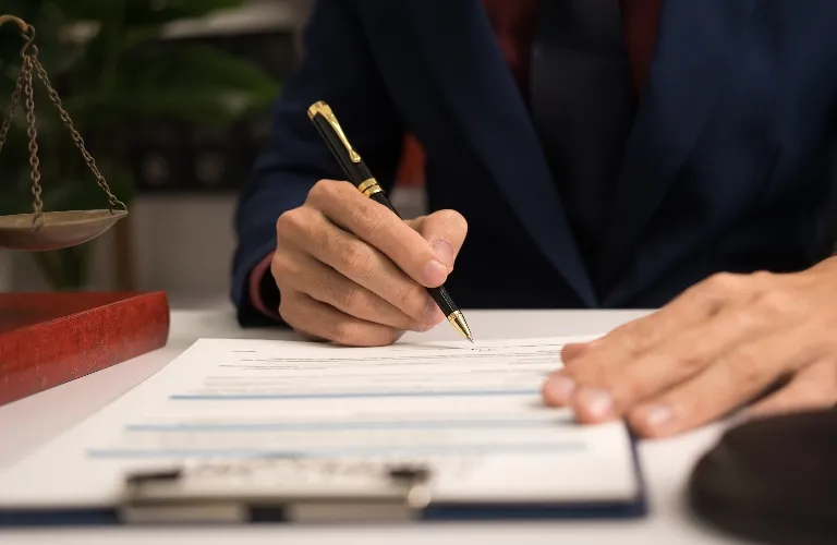 Person in dark suit signing a document on a clipboard with a pen, scales of justice nearby.