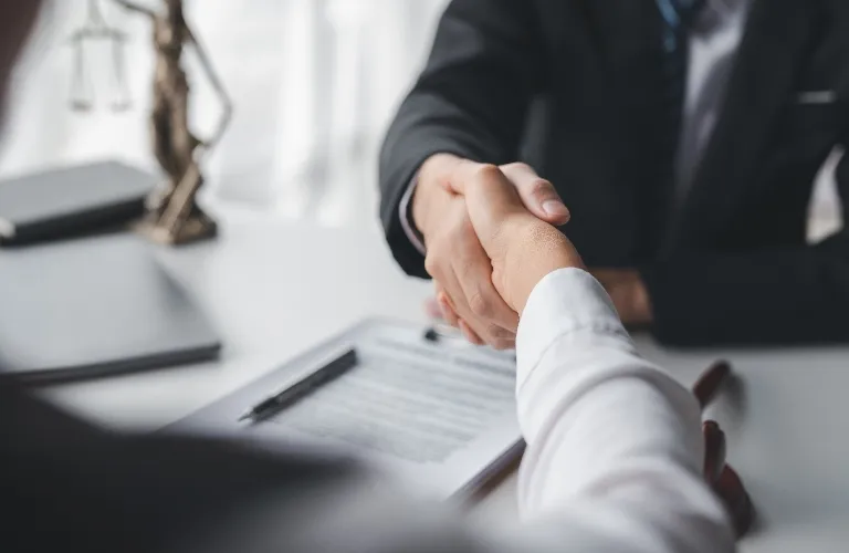 Two professionals in suits shaking hands over a desk with documents and a pen.