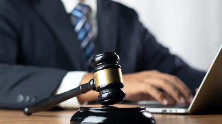 Close-up of a wooden judge's gavel on a desk with a person in a suit typing on a laptop.