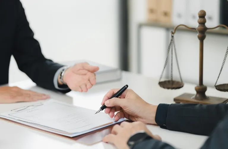 Two individuals in formal attire engage in signing and discussing documents at a white desk, with a brass balance scale in the background.