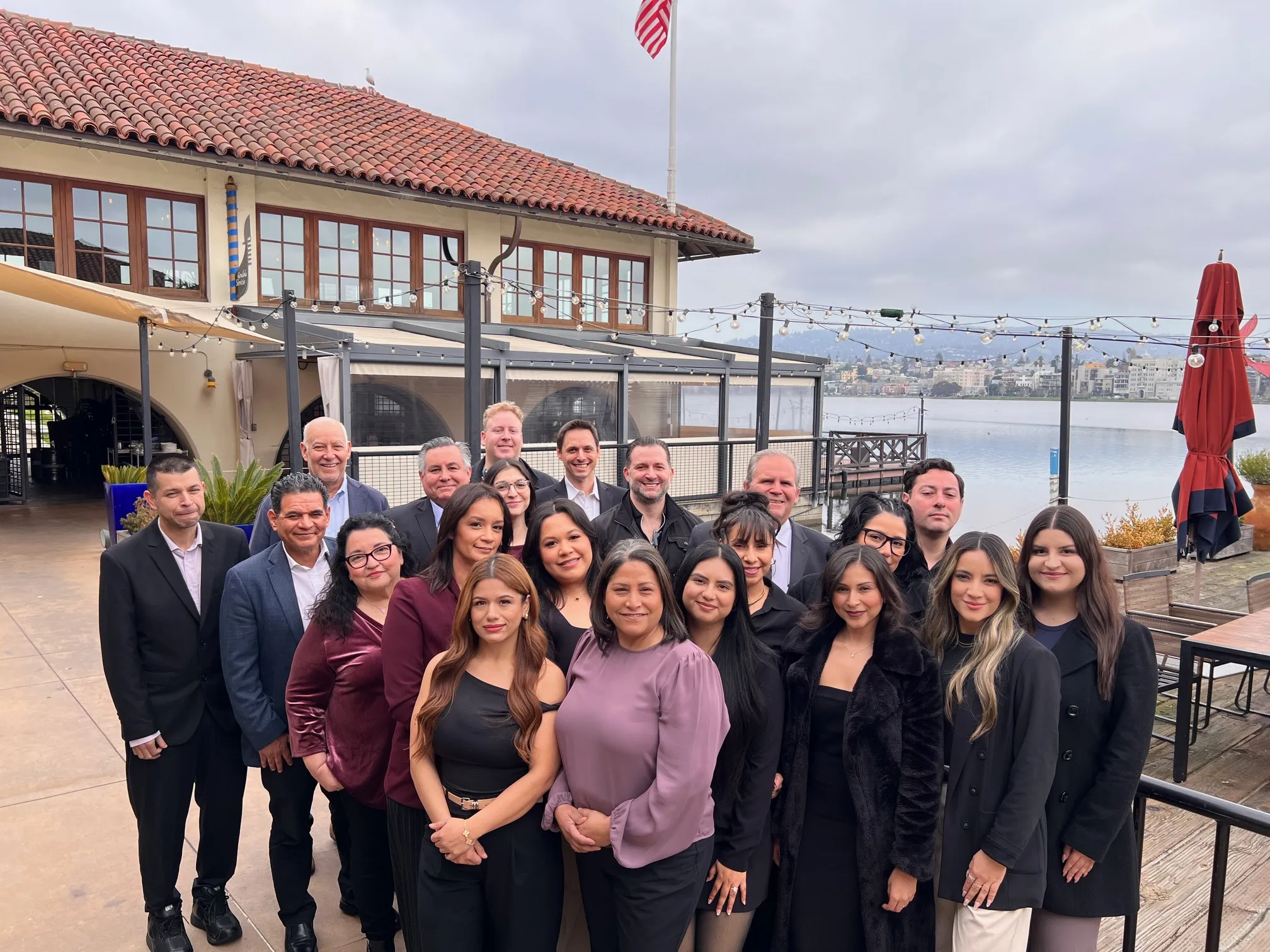 Group of professionally dressed men and women posing outdoors by waterfront building.