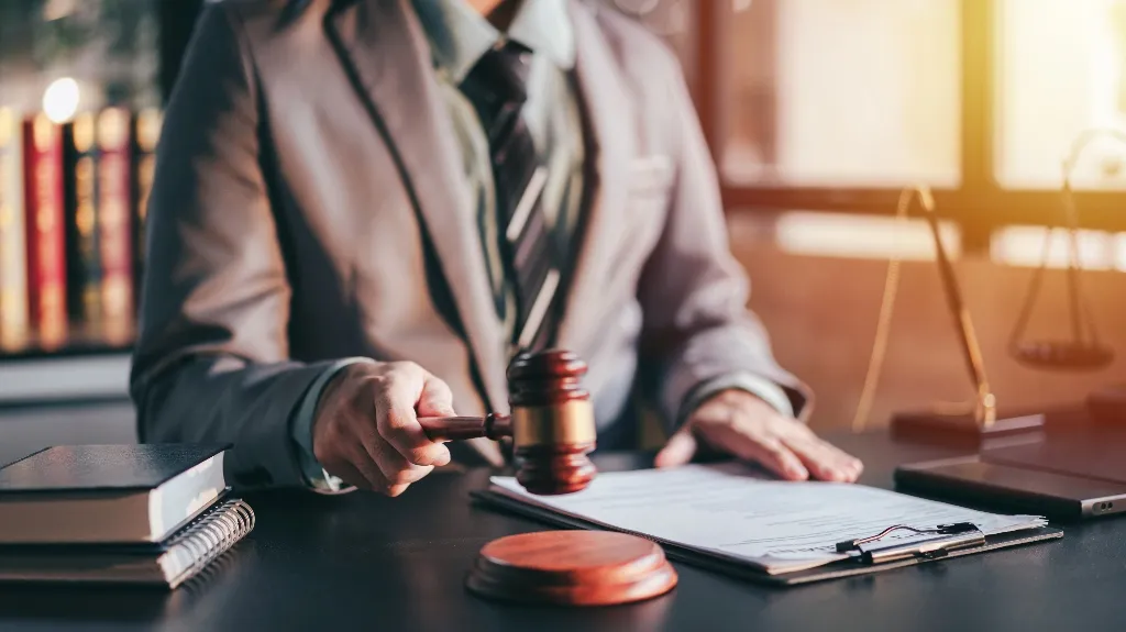 Person in suit holding wooden gavel over legal documents on desk with law books and scales.