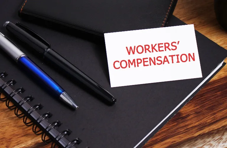 White card with red text "WORKERS' COMPENSATION" on black notebook with two pens.