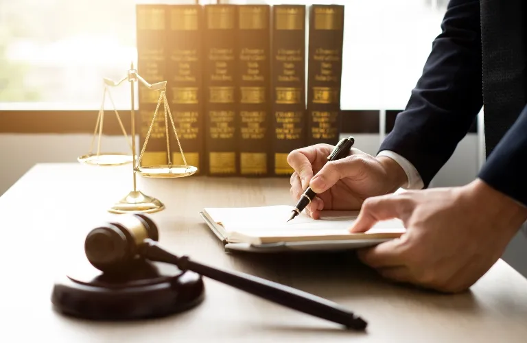 Person in a suit writing on a document at a desk with legal books, scales of justice, and a gavel.