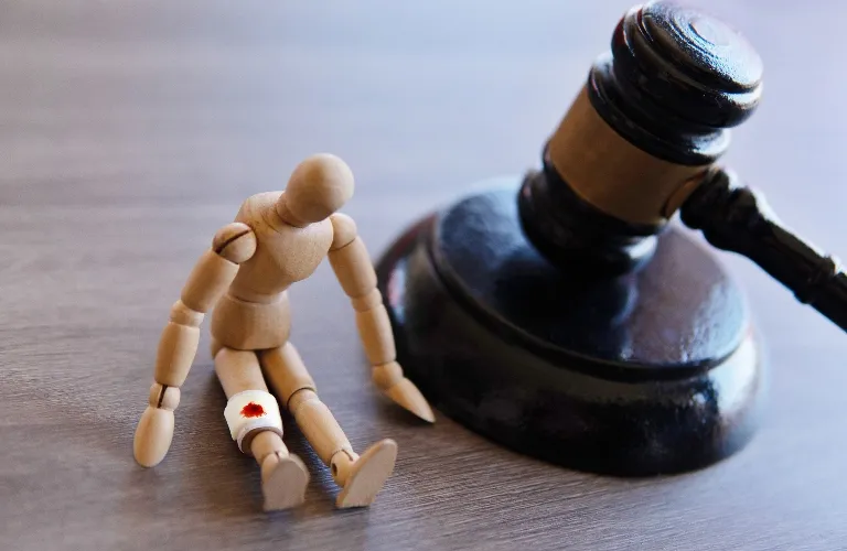 Wooden artist mannequin with a bloodied bandage on its knee seated next to a dark wooden judge's gavel.