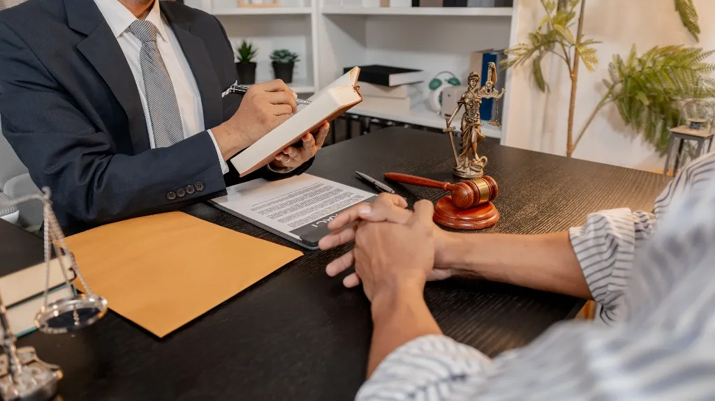 Two individuals seated at a desk with legal documents, a gavel, and Lady Justice statue.