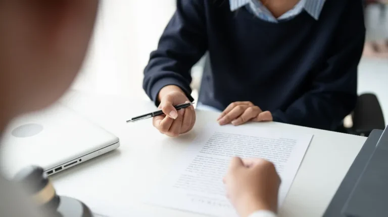 Two individuals engaged in a discussion over a document on a white desk with a laptop nearby.
