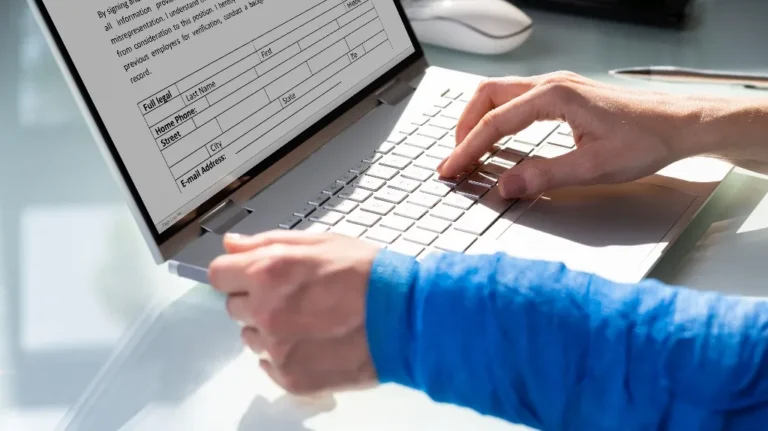 Person typing on a silver laptop displaying a form with fields for personal information.