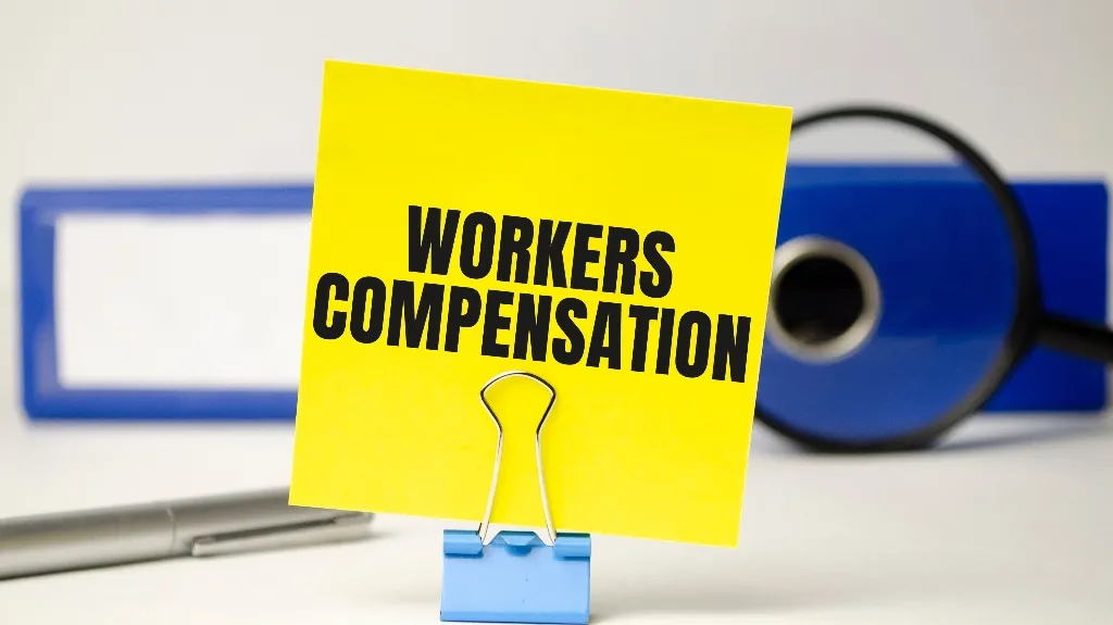 Yellow note with "WORKERS COMPENSATION" text held by blue clip, magnifying glass and blue binder in background.
