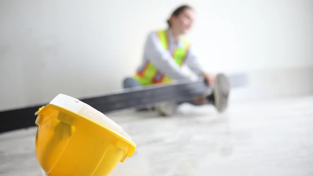 Yellow construction helmet in foreground with a blurred worker wearing a safety vest in the background.