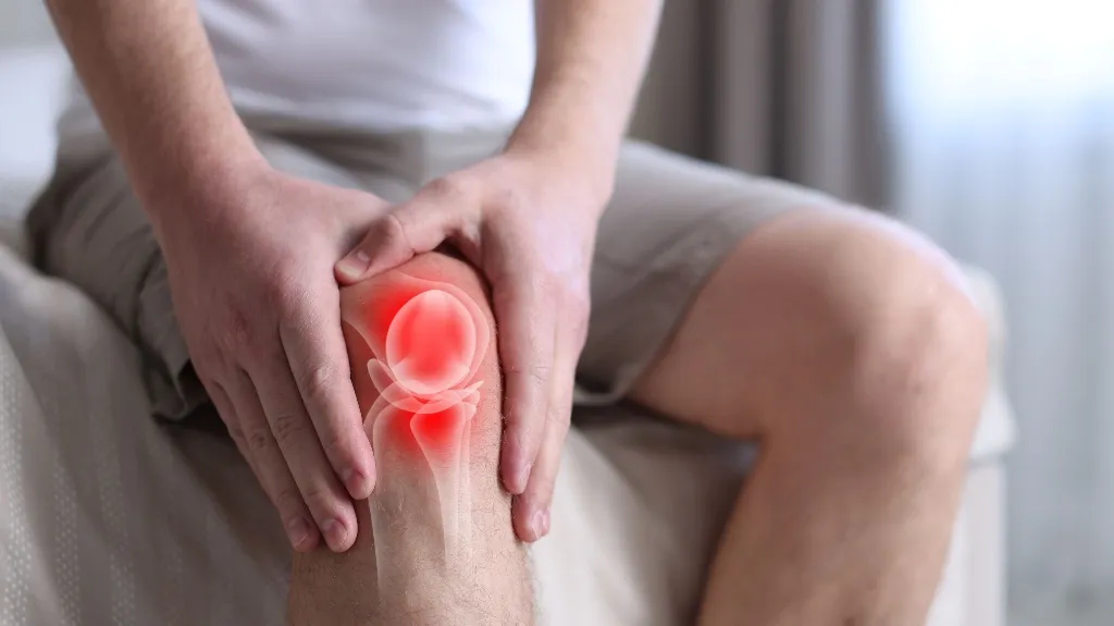 Person wearing beige shorts holding a left knee with red highlighted joint pain area.