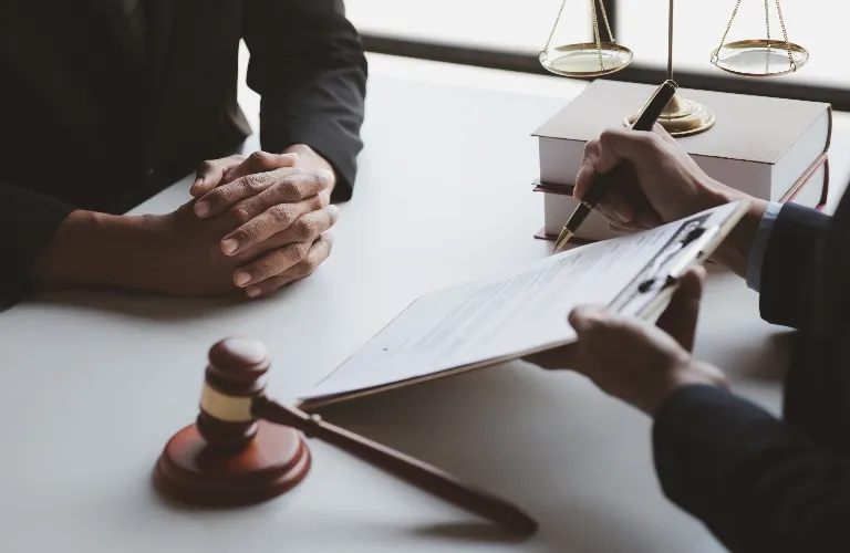 Two professionals at a desk with legal books, a gavel, and a person signing a document.