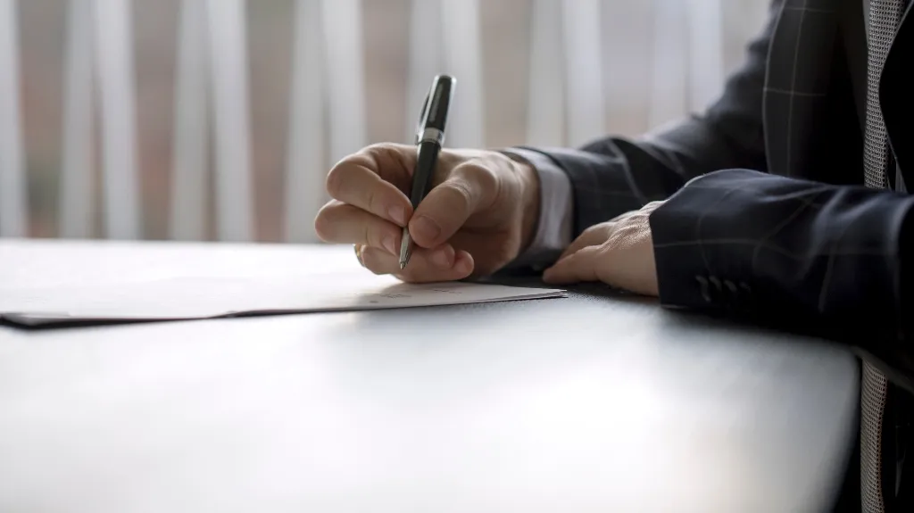 Close-up of a person in a dark suit signing a document with a black pen.