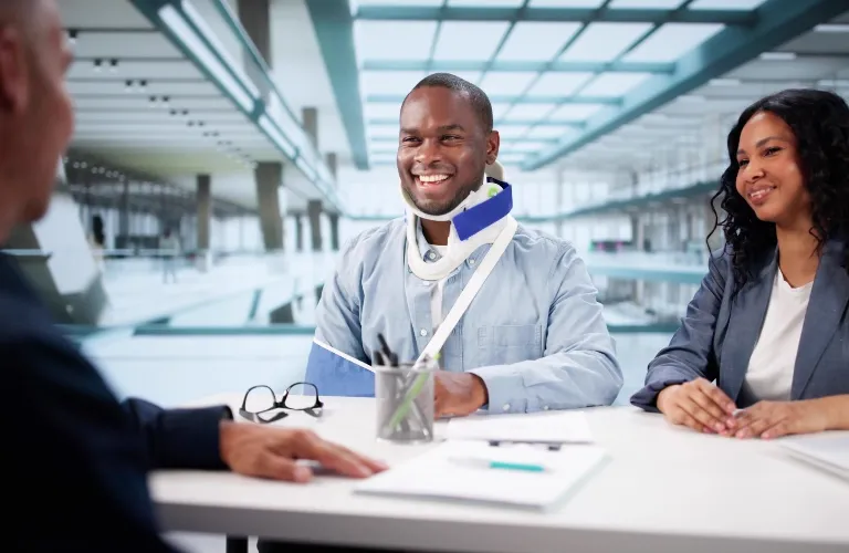 Three professionally dressed individuals engaged in a conversation at a modern office, one man wearing a neck brace and arm sling.