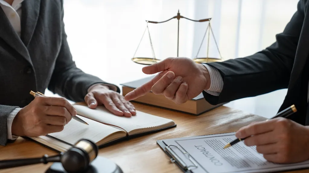Two professionals in suits discussing and signing legal documents at a wooden desk.