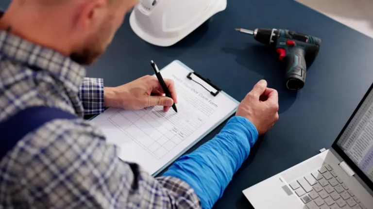 Man in plaid shirt filling out an injury insurance claim form on clipboard at desk.