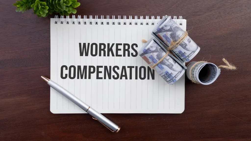 Notepad with "WORKERS COMPENSATION" text, rolled US dollar bills tied with twine, silver pen, and green plant on wooden surface.