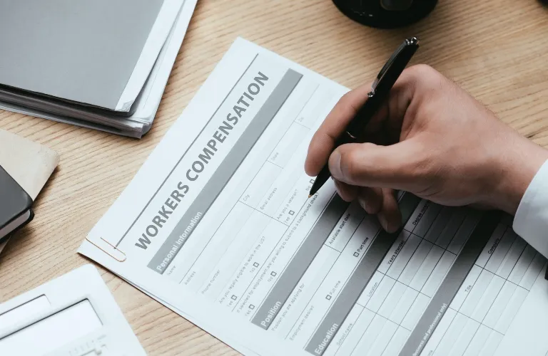 Person's hand holding a black pen filling out a workers compensation form on a wooden desk.