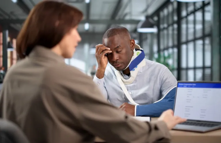 Two professionals in an office, one with a neck brace and arm sling, discussing work.
