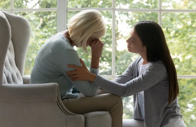 Two women indoors near a window, one comforting the other who is visibly upset.