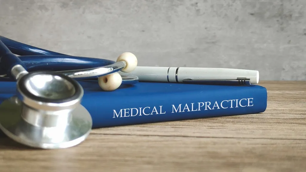 Close-up of a blue book titled "Medical Malpractice" with a stethoscope and white pen on a wooden surface.