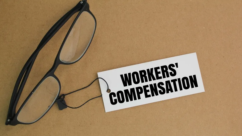 Black-framed eyeglasses with a white tag labeled "WORKERS' COMPENSATION" on a brown surface.