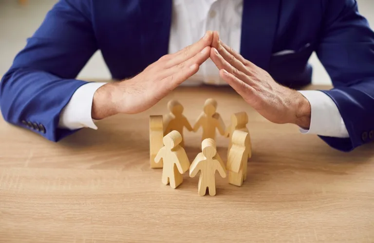 Person in blue suit forming a protective gesture over wooden human figures on a table.