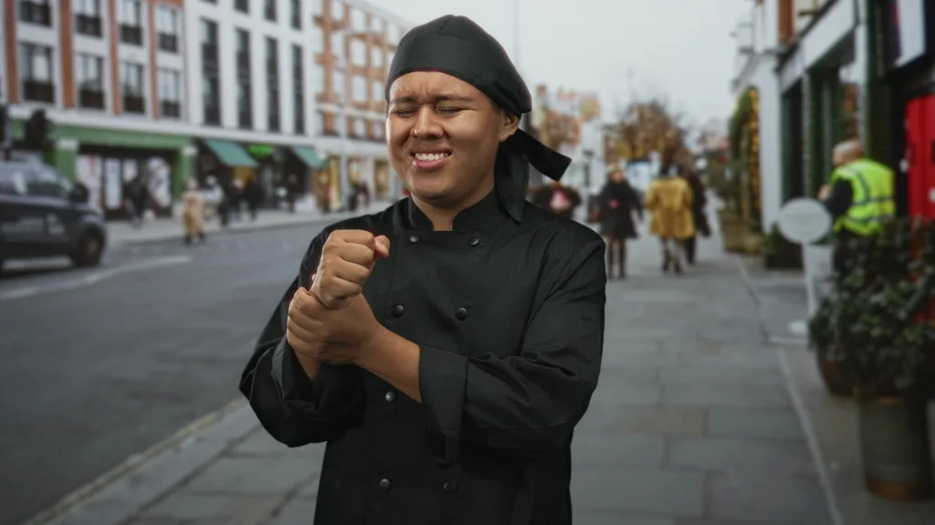 Chef in black uniform and headscarf grimacing while holding wrist on city street.