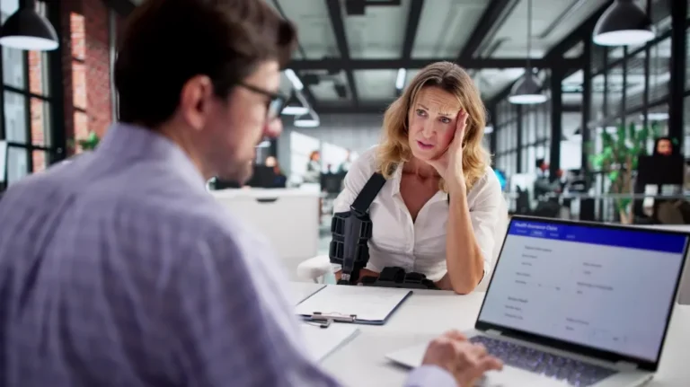 Woman with arm sling consulting a man in glasses at modern office desk with laptop.