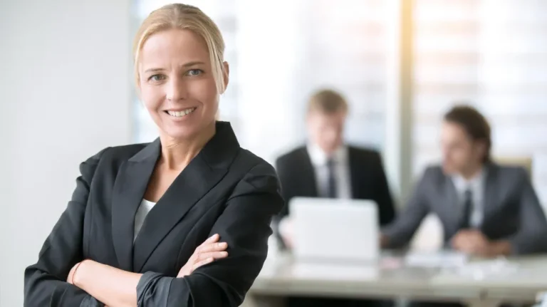Confident blonde businesswoman in black blazer with crossed arms, two men in suits blurred in background.