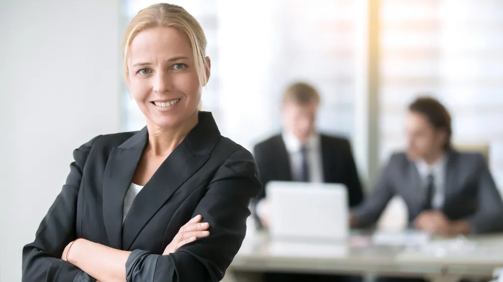 Confident blonde businesswoman in black blazer with crossed arms, two men in suits blurred in background.