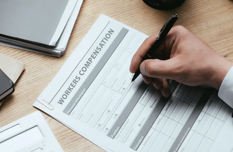 Person filling out a workers compensation form on a wooden desk with office supplies.