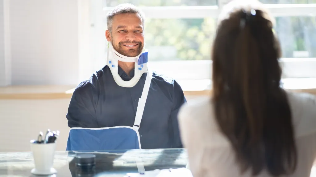 Smiling man wearing neck brace and arm sling sitting across from woman at desk.