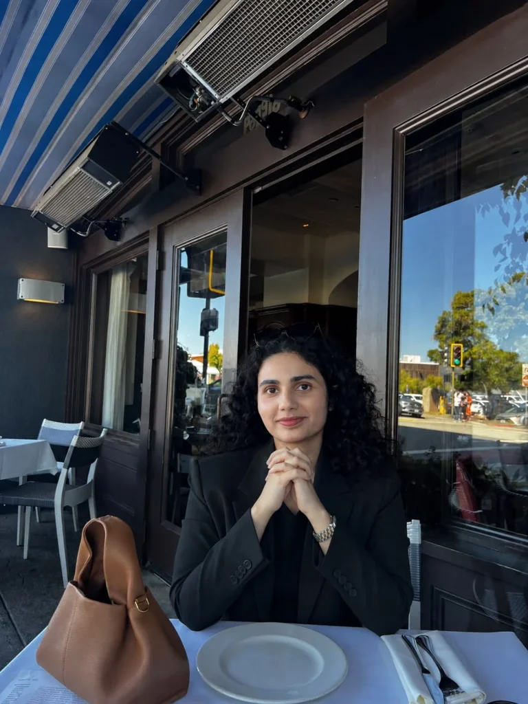 Woman with curly dark hair wearing a black blazer seated at a restaurant table with a brown handbag and white table setting.