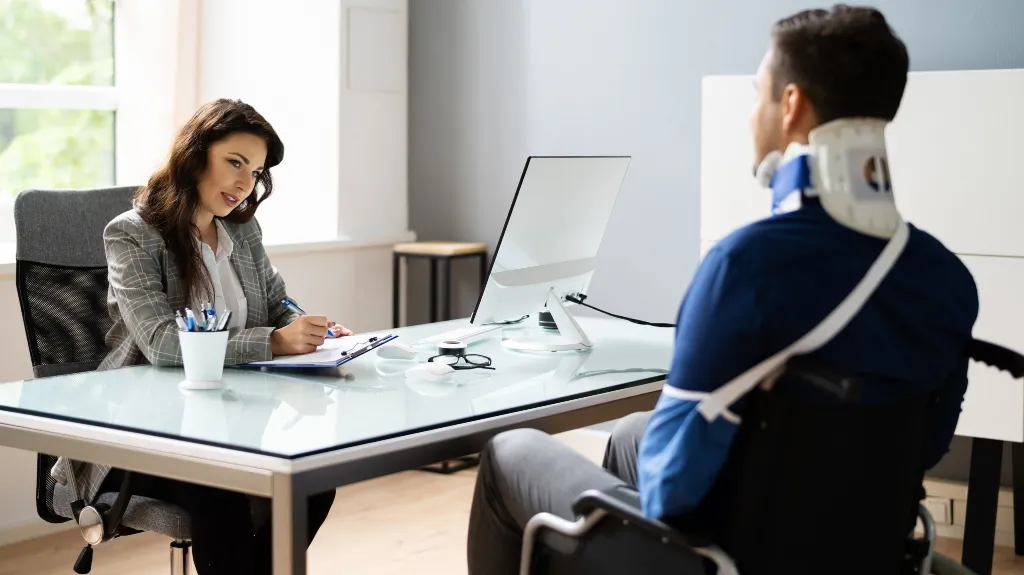 Professional woman in gray blazer interviewing man with neck brace and arm sling in office.