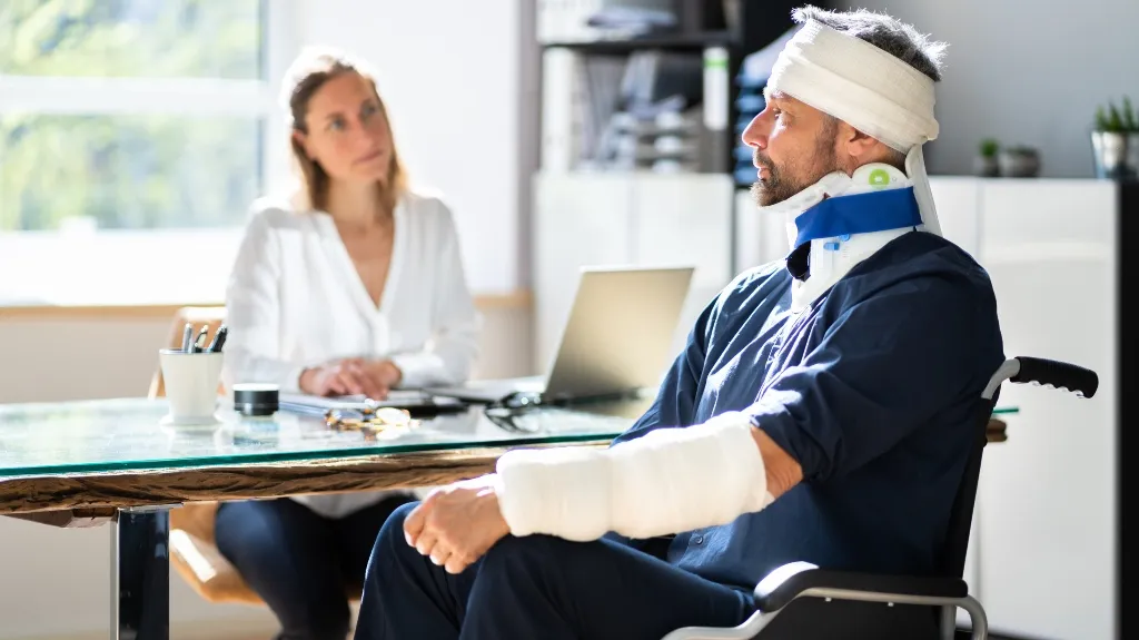 Man with head and arm bandages and neck brace sitting in wheelchair, woman in white blouse seated at desk.