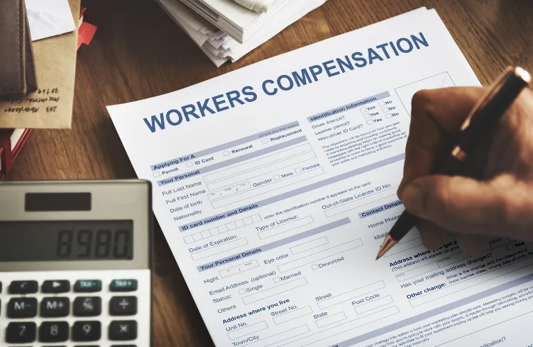Hand filling out a workers compensation form on a wooden desk with calculator.