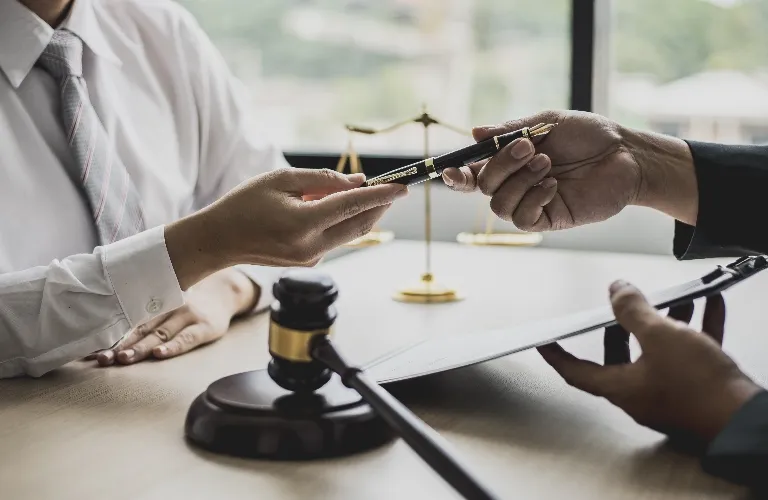 Two professionals exchanging a pen over a desk with a gavel and scales of justice.