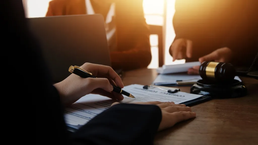 Close-up of hands signing a contract on a clipboard, with a gavel and laptop on a wooden table.